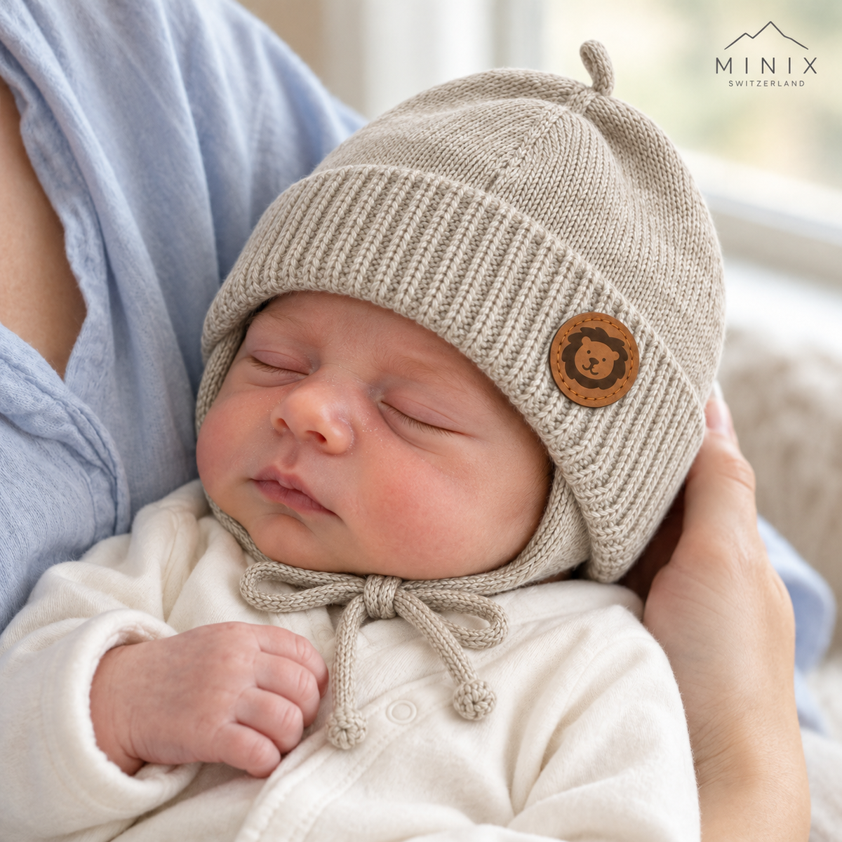 Newborn baby in a parent's arms wearing the beige MINIX knitted cap with lion patch