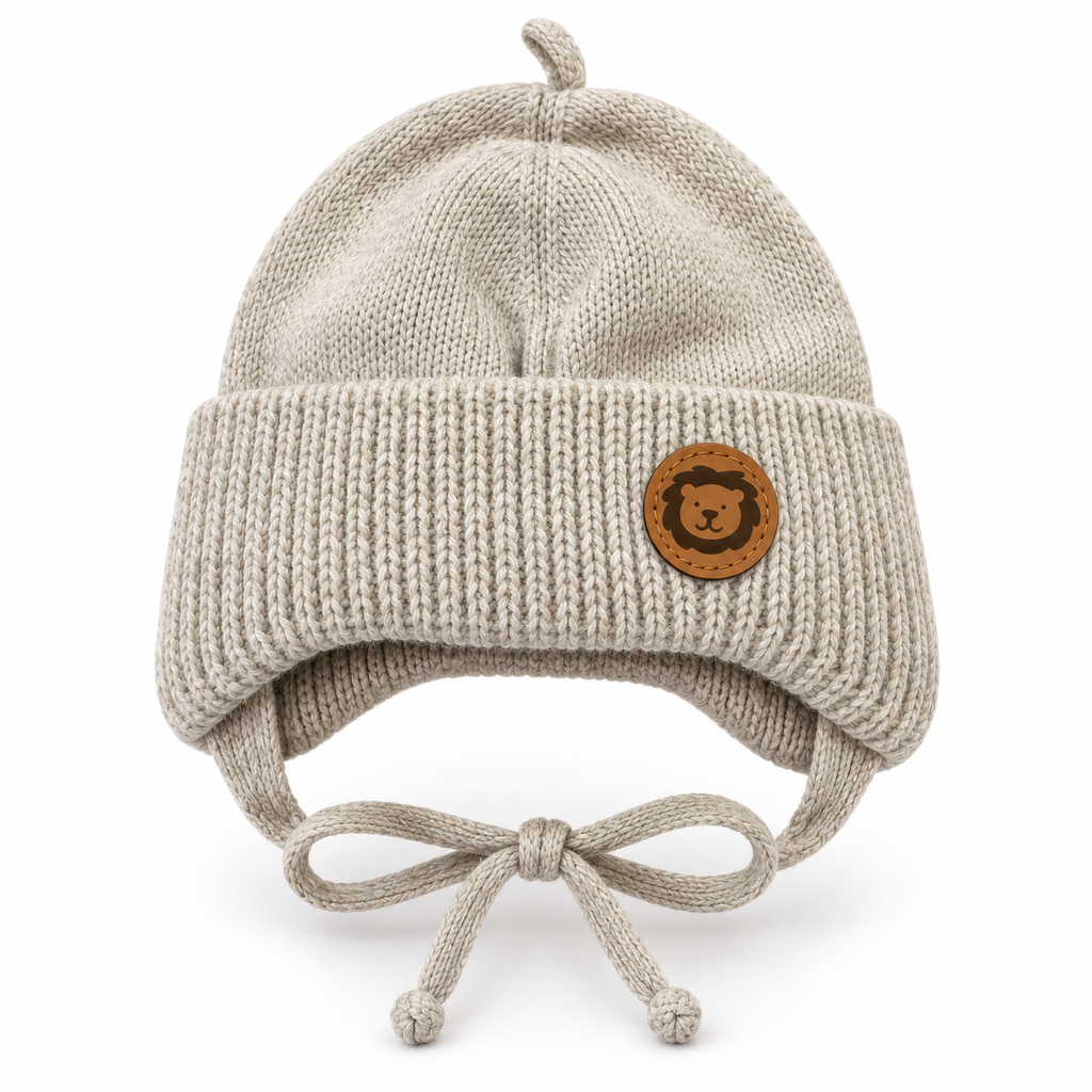 Beige knitted baby cap with lion patch, chunky ribbed cuff, ear flaps and tie strings – front view