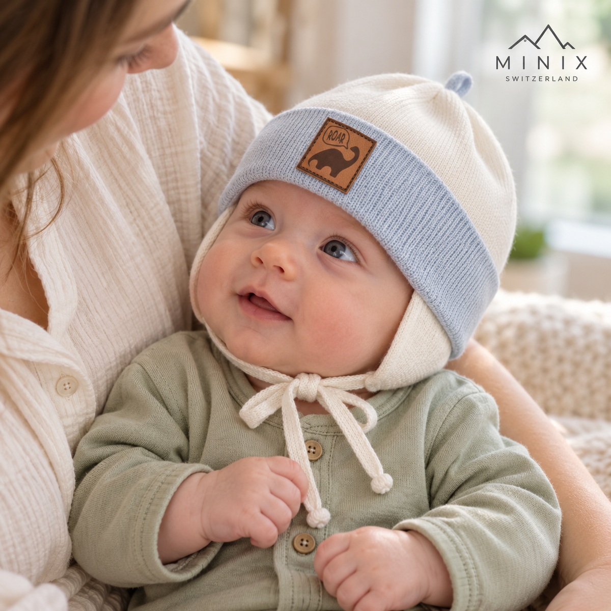 Smiling baby in his mother's arms wearing the cream and light blue MINIX knitted dino cap