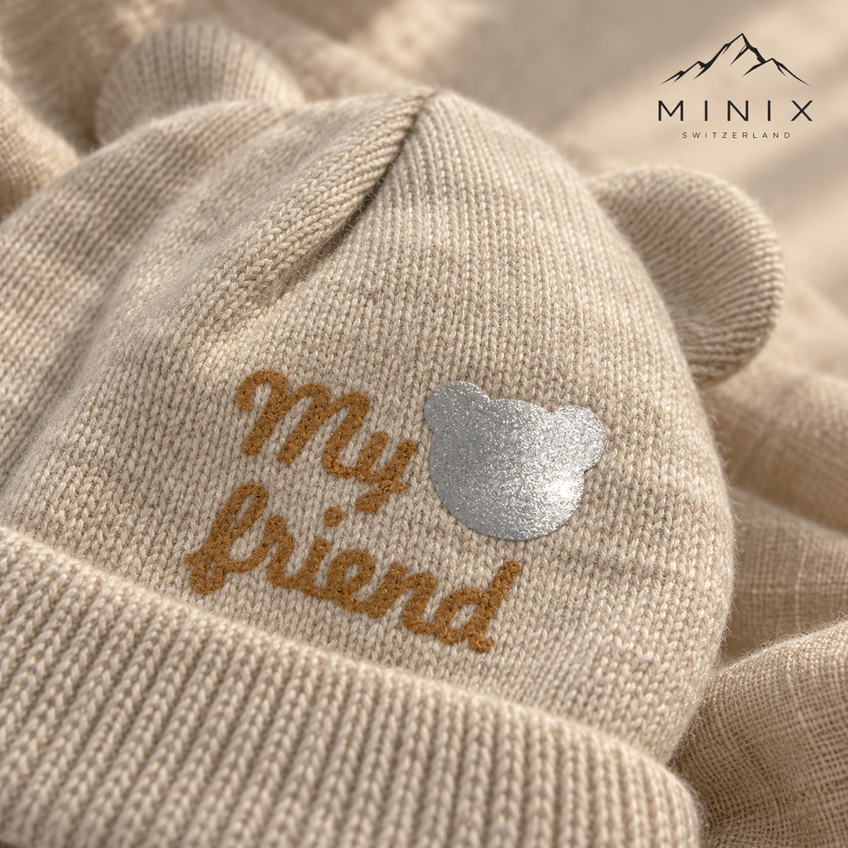 Close-up of the "My friend" lettering and silver bear patch on the beige knitted cap