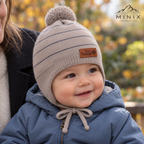 Smiling baby boy held by a parent, wearing the beige MINIX striped knitted pompom cap