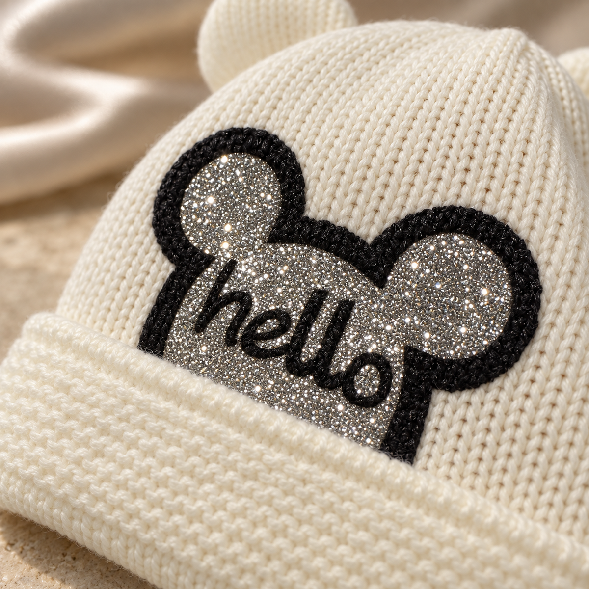Close-up of the sparkly glitter hello patch on a cream knitted baby cap