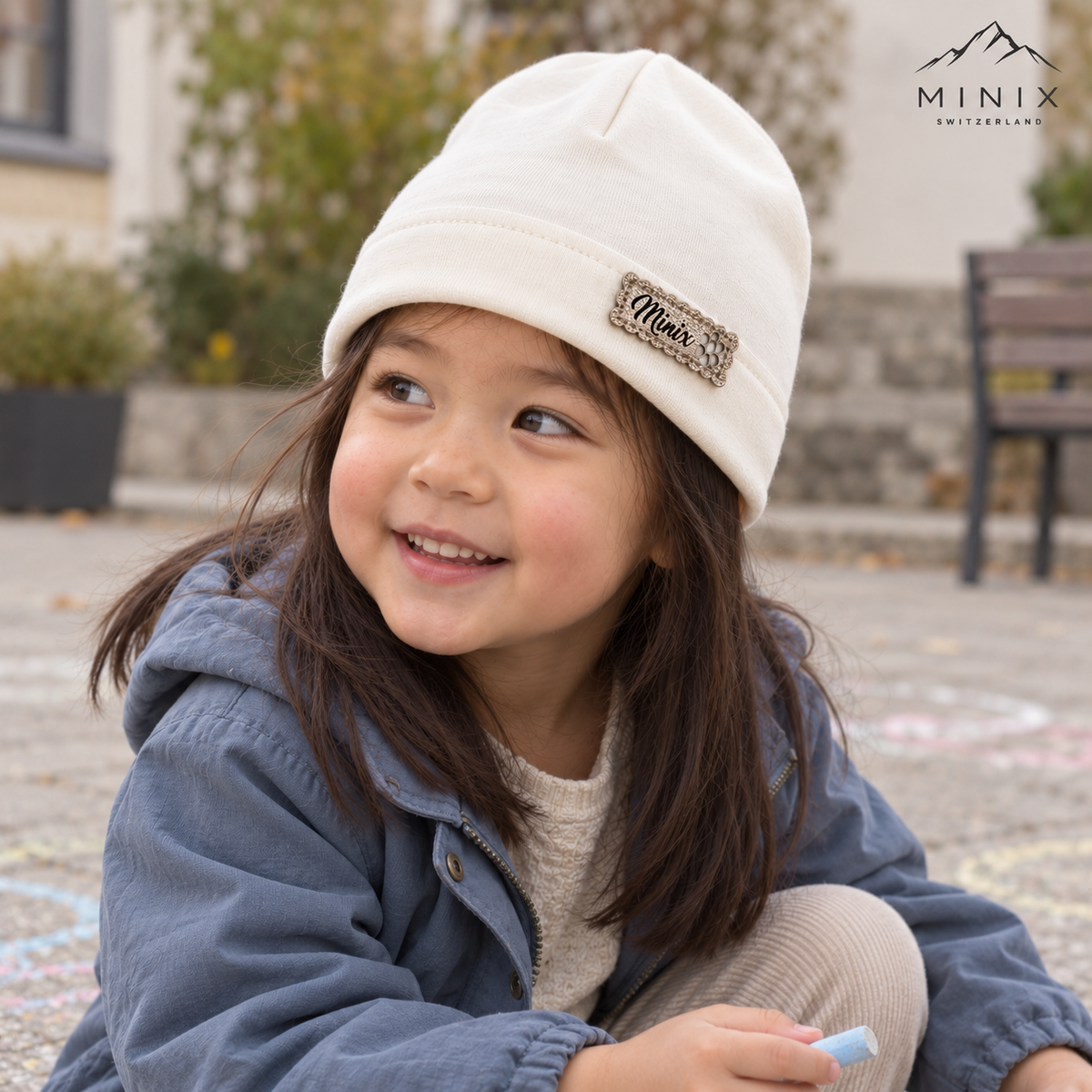 Smiling girl playing with chalk outdoors, wearing a cream MINIX cotton beanie
