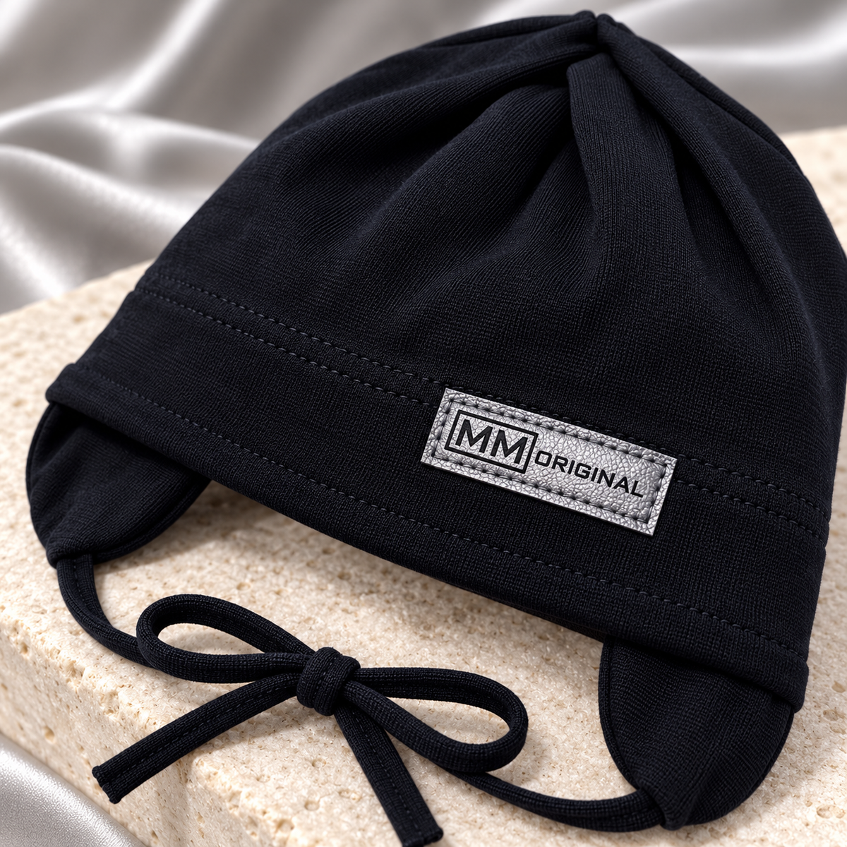 Close-up of the silver MM Original label on a black kids cotton cap with ear flaps and ties